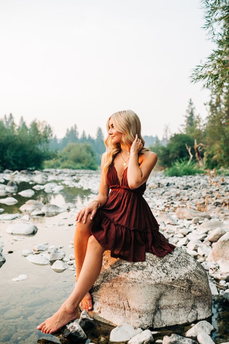 a woman sitting on top of a rock next to a river wearing a red dress