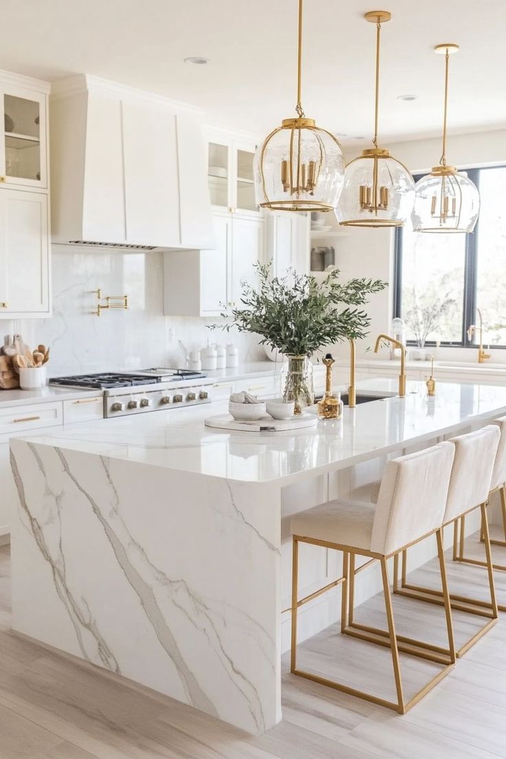 a white kitchen with marble counter tops and gold accents on the island in front of two windows