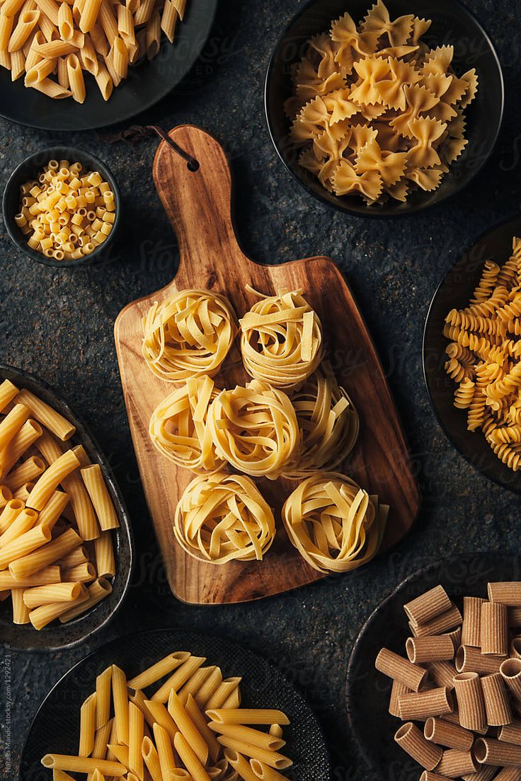 various types of pasta in bowls on a table next to wooden spoons and cutting board