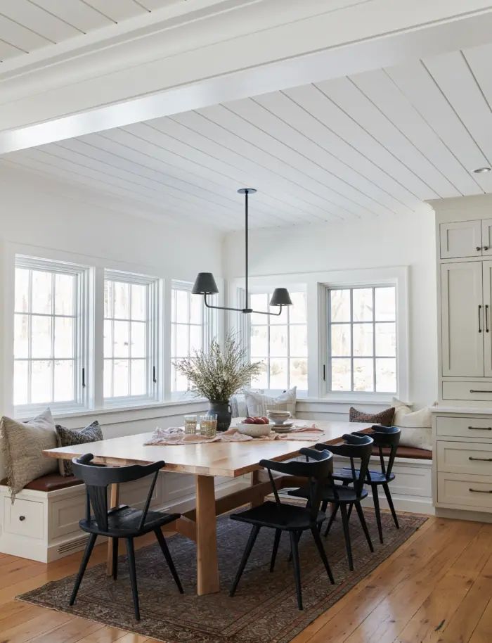 a dining room table with chairs and a bench in front of two windows on the wall