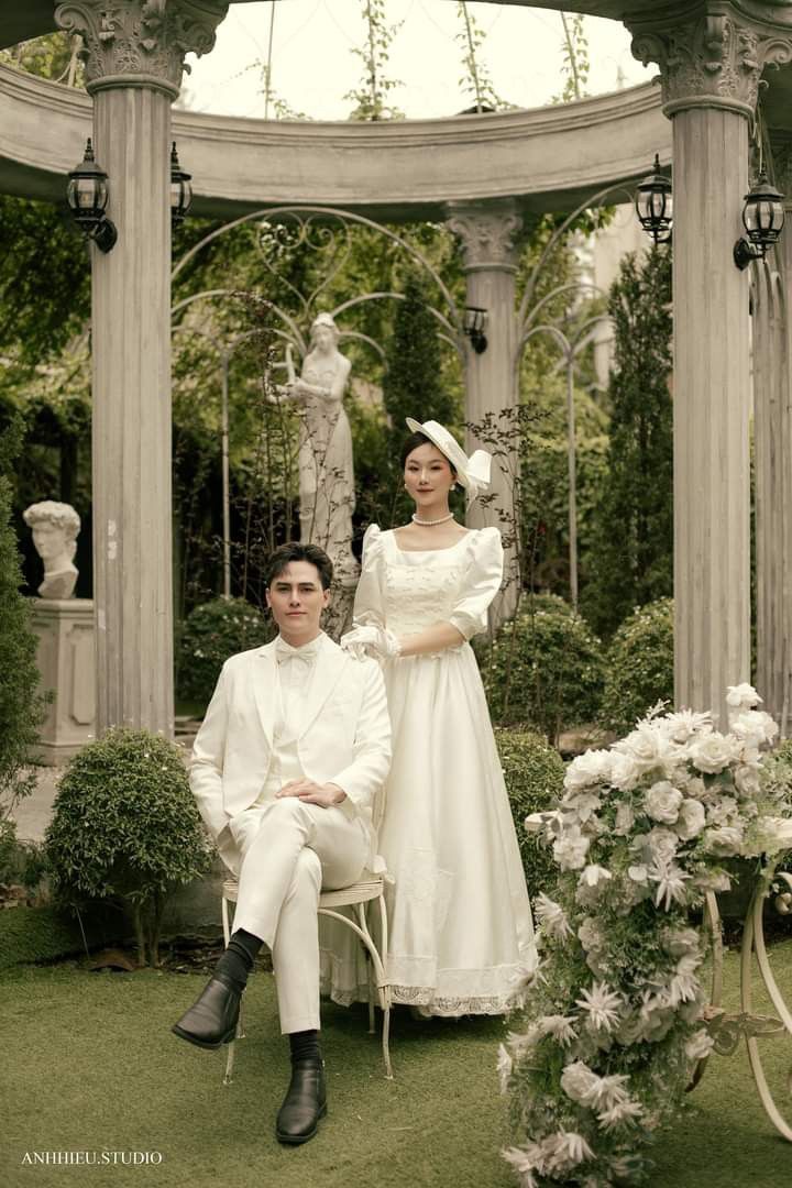 a man and woman dressed in period clothing sitting on lawn chairs under an arbor with white flowers