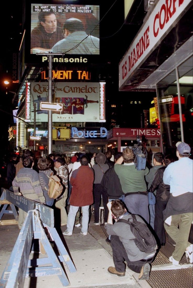 a crowd of people standing on the side of a street next to tall buildings at night