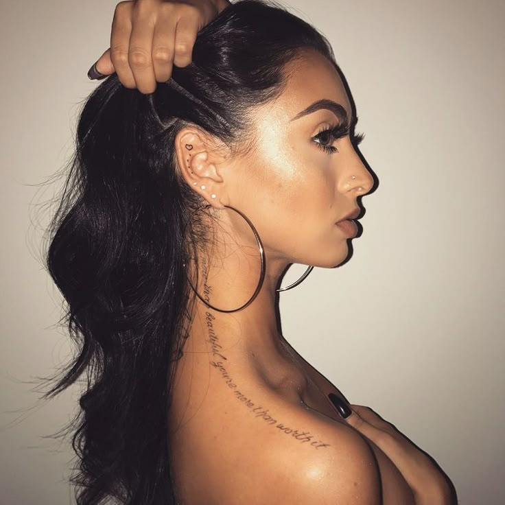 a woman with tattoos on her back and shoulder is shown from the neck down, she has long dark hair and wears large hoop earrings