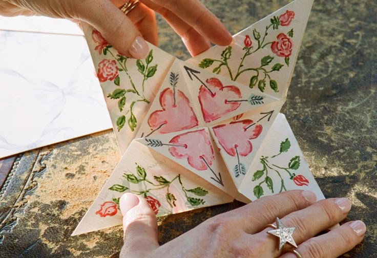 two hands are holding origami pieces with flowers on them and one hand is reaching for the paper