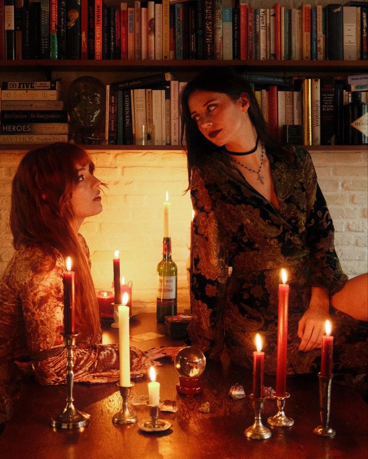 two women sitting at a table with candles in front of them and bookshelves behind them