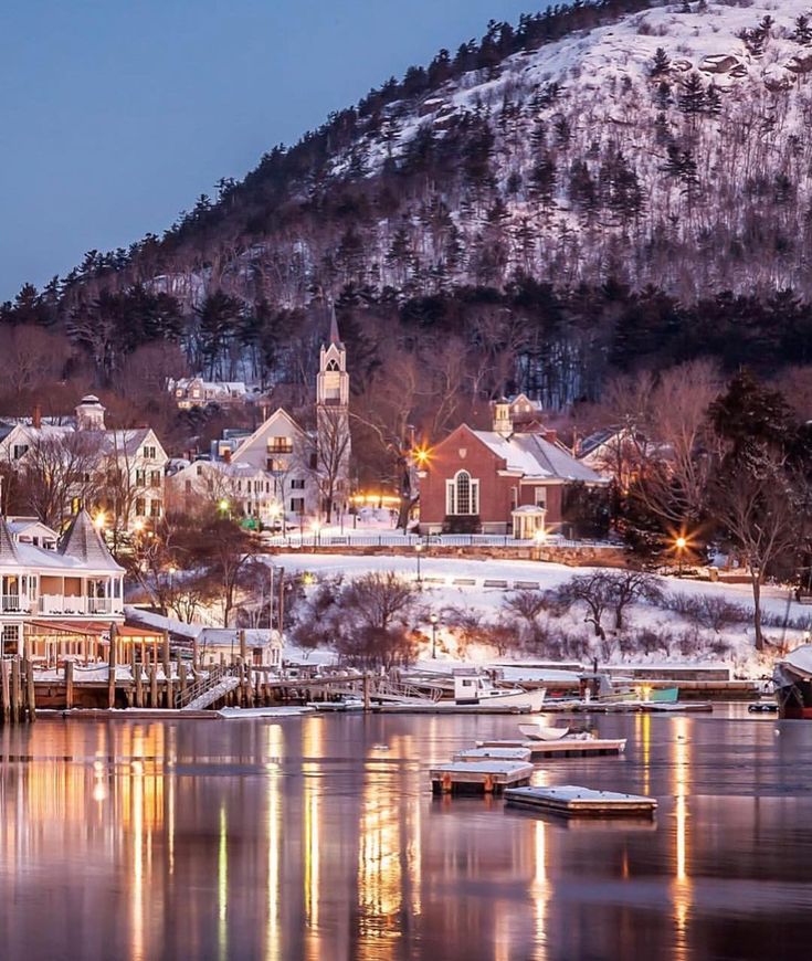 the town is lit up at night by the water with snow on the mountains in the background