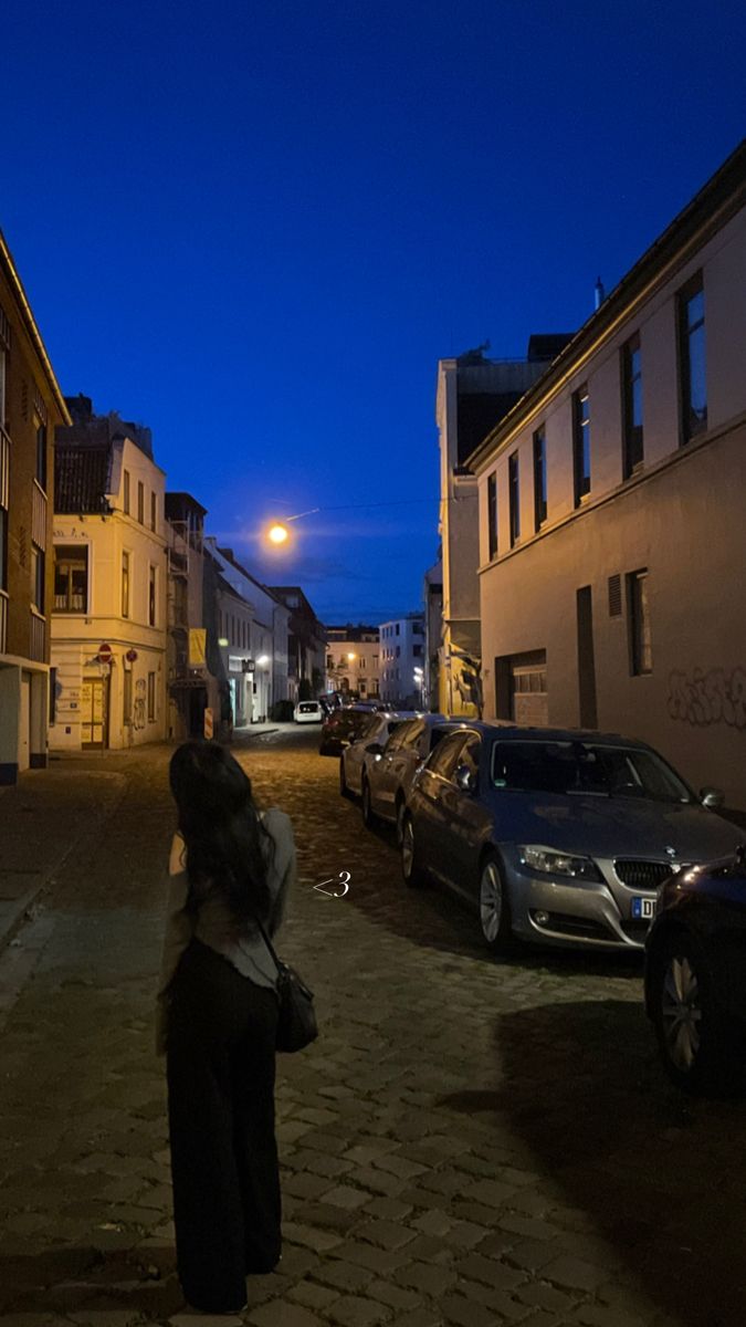 a woman standing on the side of a road next to parked cars and buildings at night