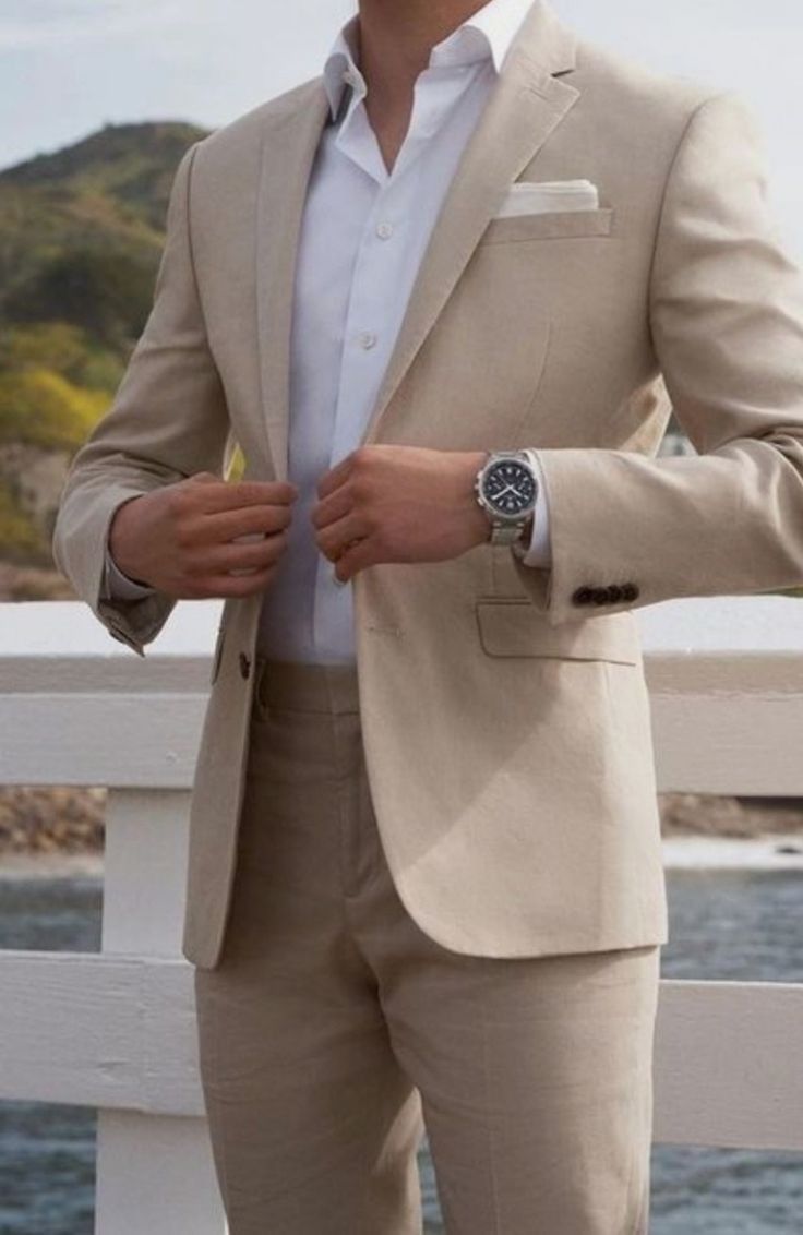 a man in a tan suit and white shirt is standing on a bridge by the water