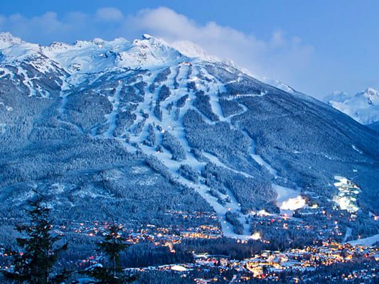 a snowy mountain covered in lots of snow next to a town and ski slope at night