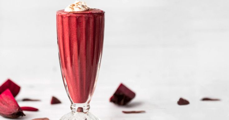 a glass filled with red liquid next to pieces of chocolate and rose petals on a white surface