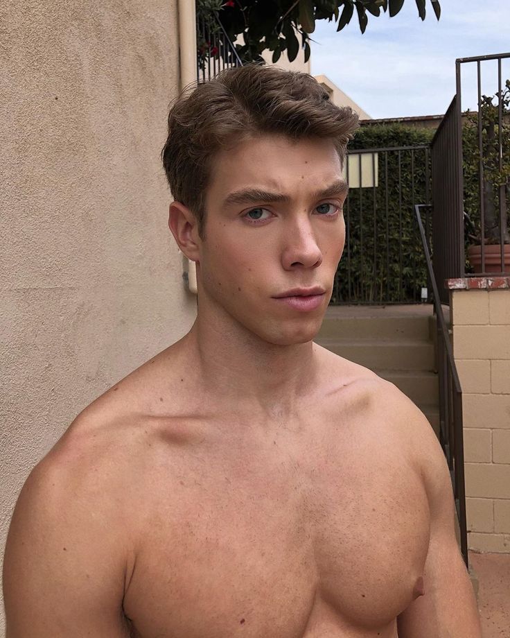 a shirtless young man standing in front of a building with his hand on his hip