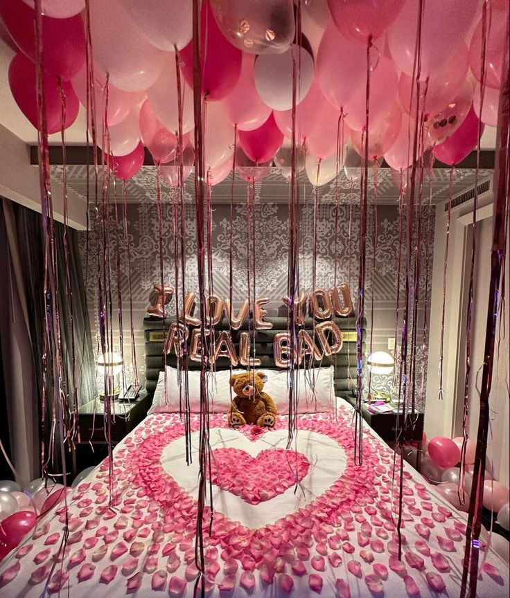 a teddy bear sitting on top of a bed surrounded by pink balloons and streamers