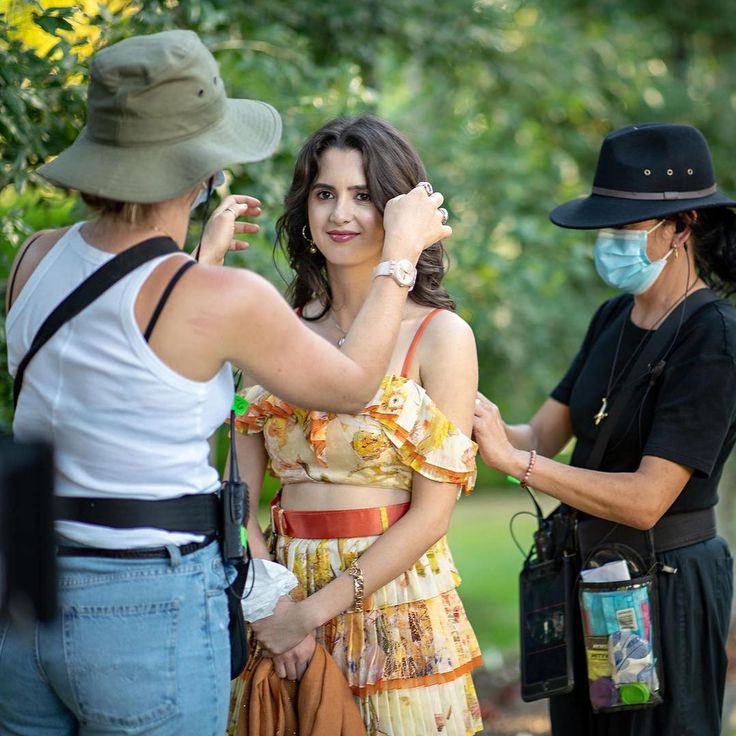 a woman wearing a face mask while standing next to another woman in a dress and hat