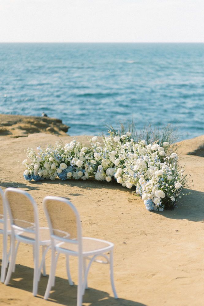 two white chairs sitting on top of a beach next to the ocean with flowers growing out of them