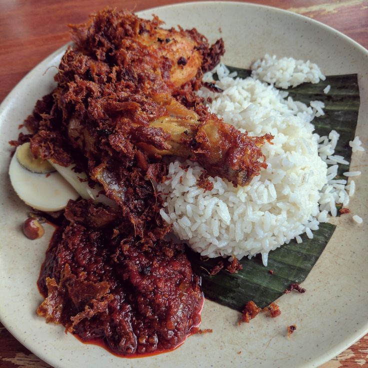 a white plate topped with meat and rice on top of a wooden table next to a banana leaf