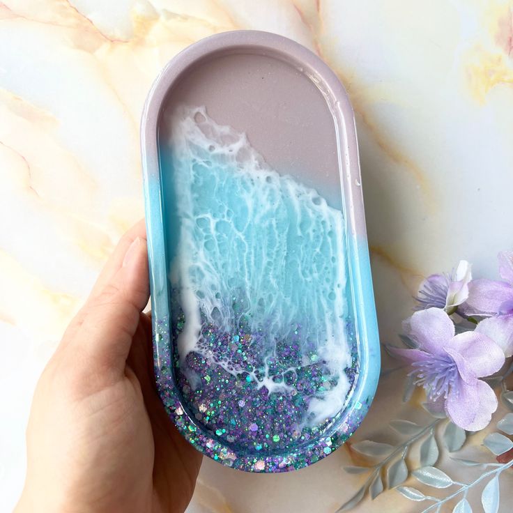 a hand holding a blue and purple soap dish with water in it next to flowers