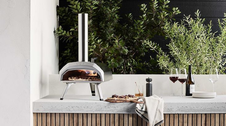 an outdoor pizza oven sitting on top of a counter next to wine glasses and bottles