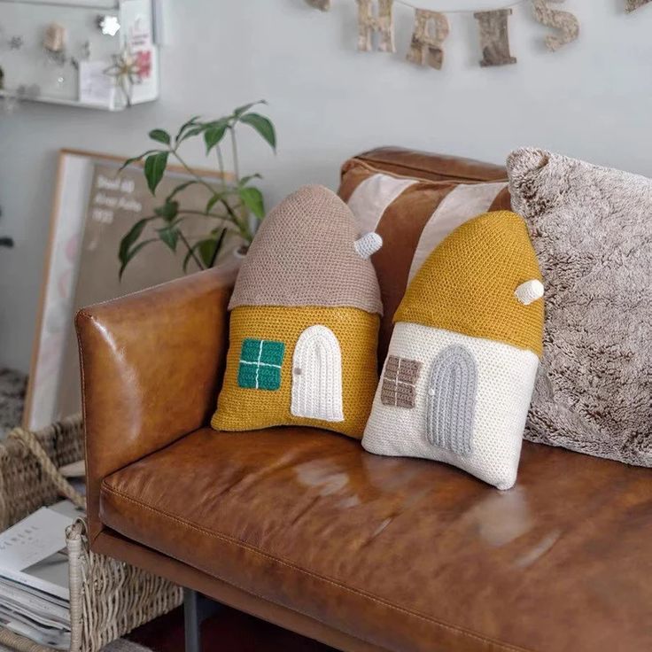 two knitted house pillows sitting on top of a brown leather couch next to a potted plant