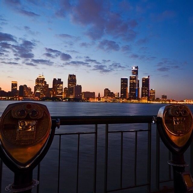 there are two binoculars that are on the railing by the water and city lights in the background