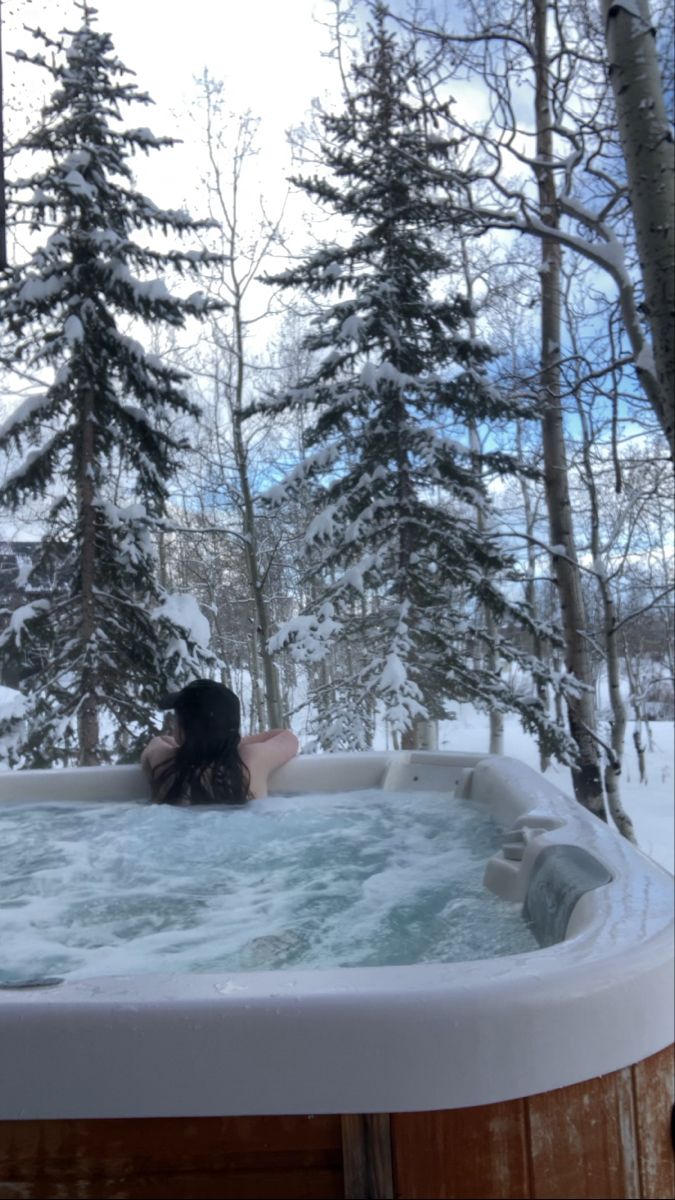 a woman laying in a hot tub surrounded by snow covered trees and evergreens on a snowy day