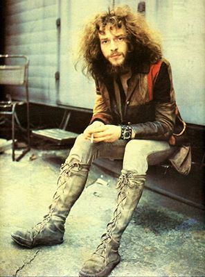 a man with long hair and boots sitting on a bench in front of a building