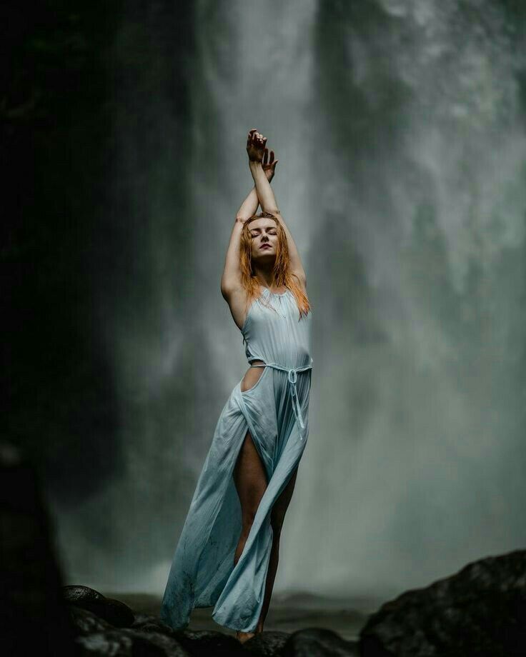 a woman standing in front of a waterfall with her hands up to the sky,