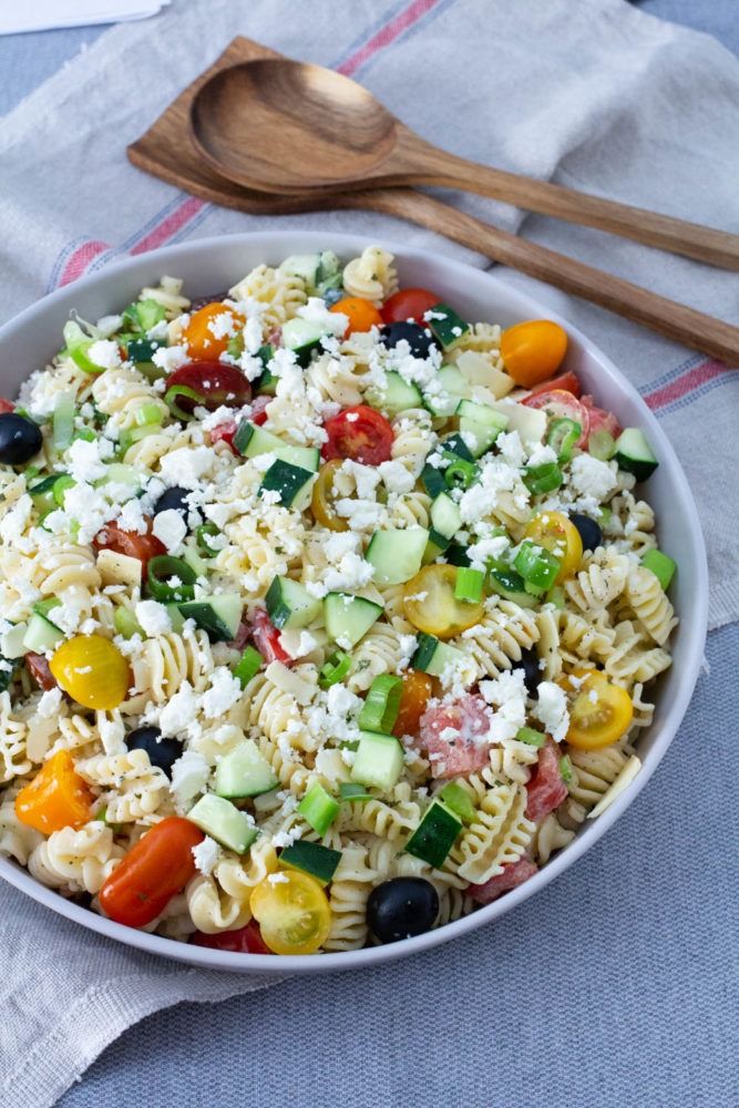 a white bowl filled with pasta salad next to a wooden spoon on top of a table