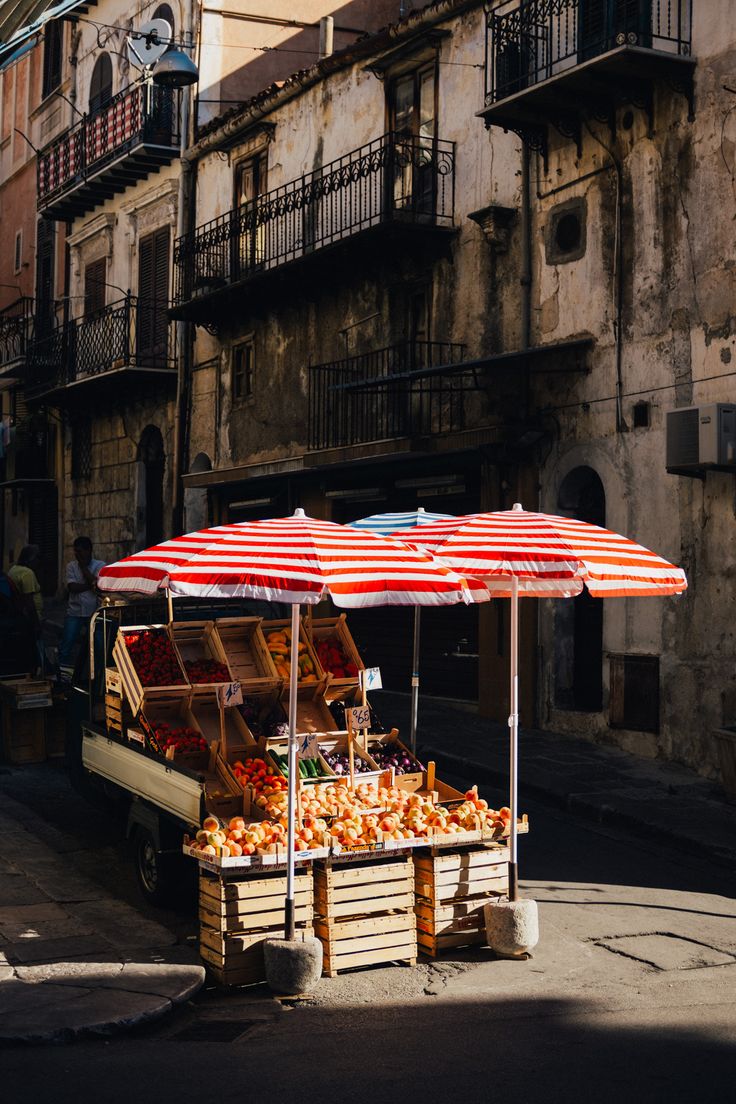 an open air fruit and vegetable stand on the side of a street with buildings in the background