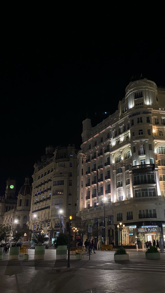 the building is lit up at night with people walking on the sidewalk in front of it