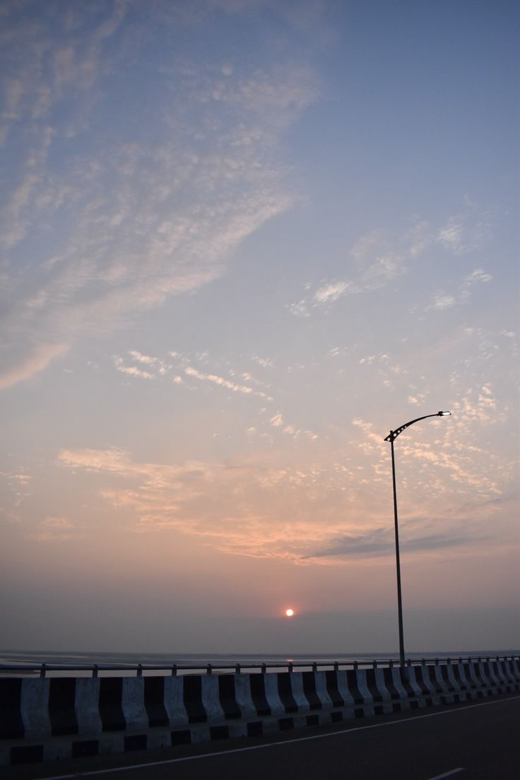 the sun is setting over the ocean with clouds in the sky and street lights on either side