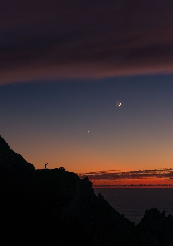 a person standing on top of a mountain at sunset with the moon in the distance