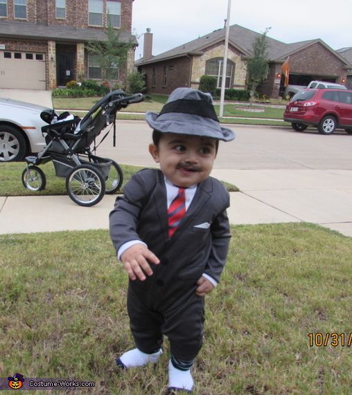 a baby in a suit and tie standing on the grass with a stroller behind him