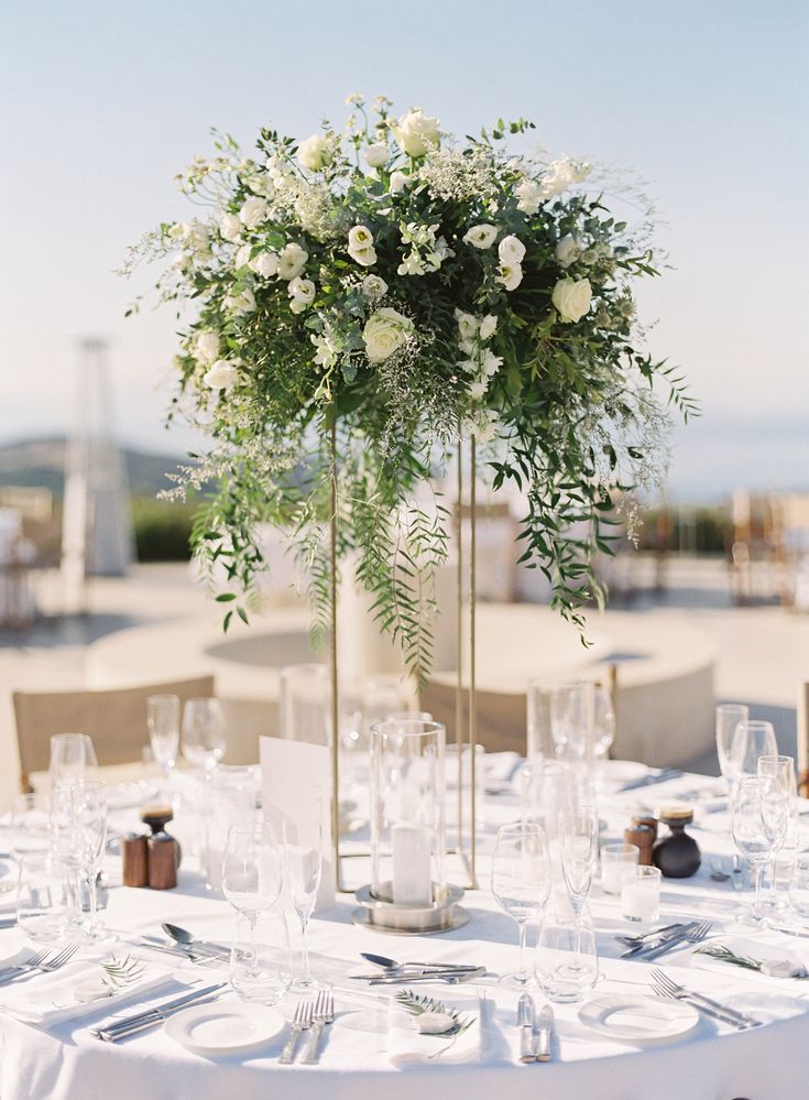 a tall vase filled with white flowers and greenery on top of a round table