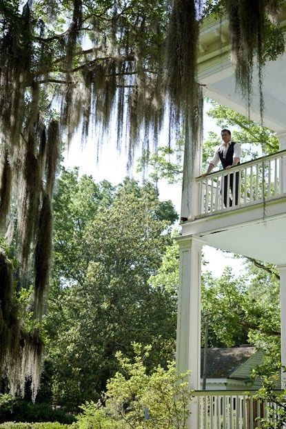 a man standing on the balcony of a white house with moss hanging from it's ceiling