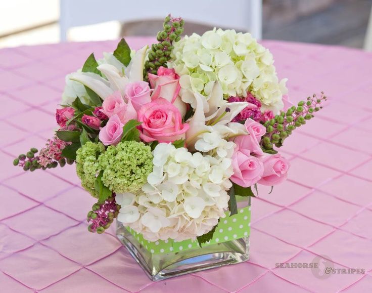 a square vase filled with white and pink flowers on top of a purple table cloth