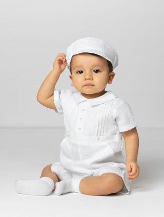 a baby sitting on the floor wearing a white outfit and holding his hand up to his head