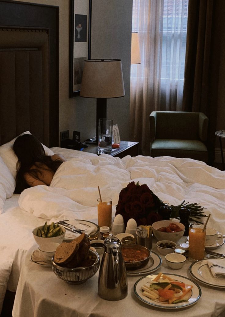 a woman is laying in bed with her head on the table full of food and drinks