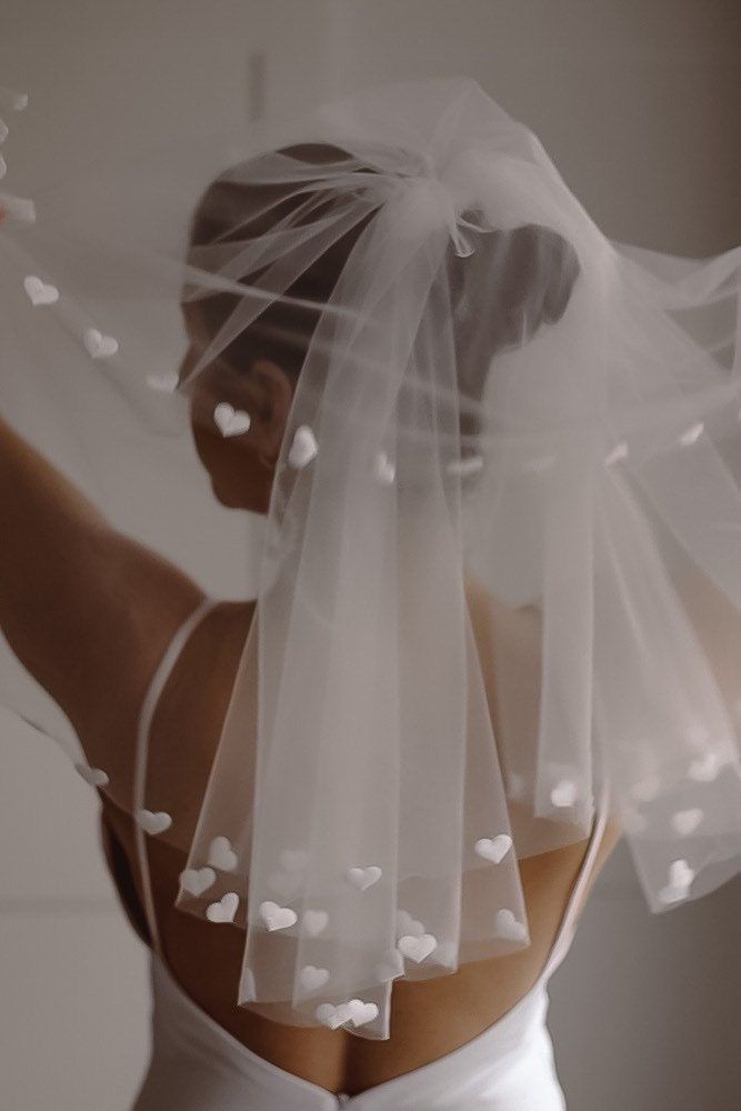 the back of a bride's wedding dress with white hearts on it and her veil blowing in the wind