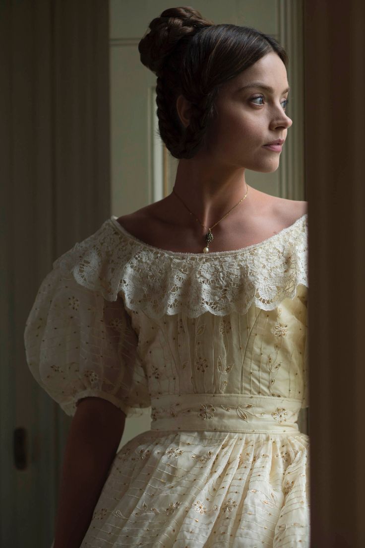 a woman in a white dress looking out the window with her hand on her hip