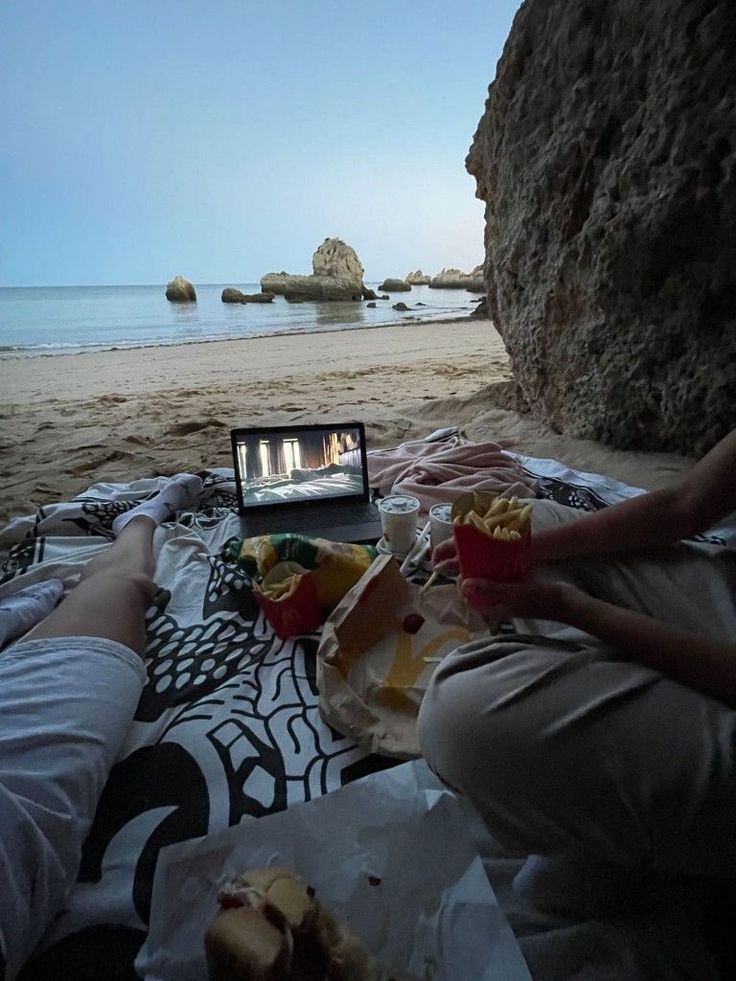 two people sitting on the beach with food and drinks in front of them, watching tv