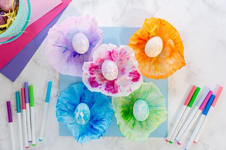 colorful paper flowers and pens on a marble counter top with easter eggs in the center