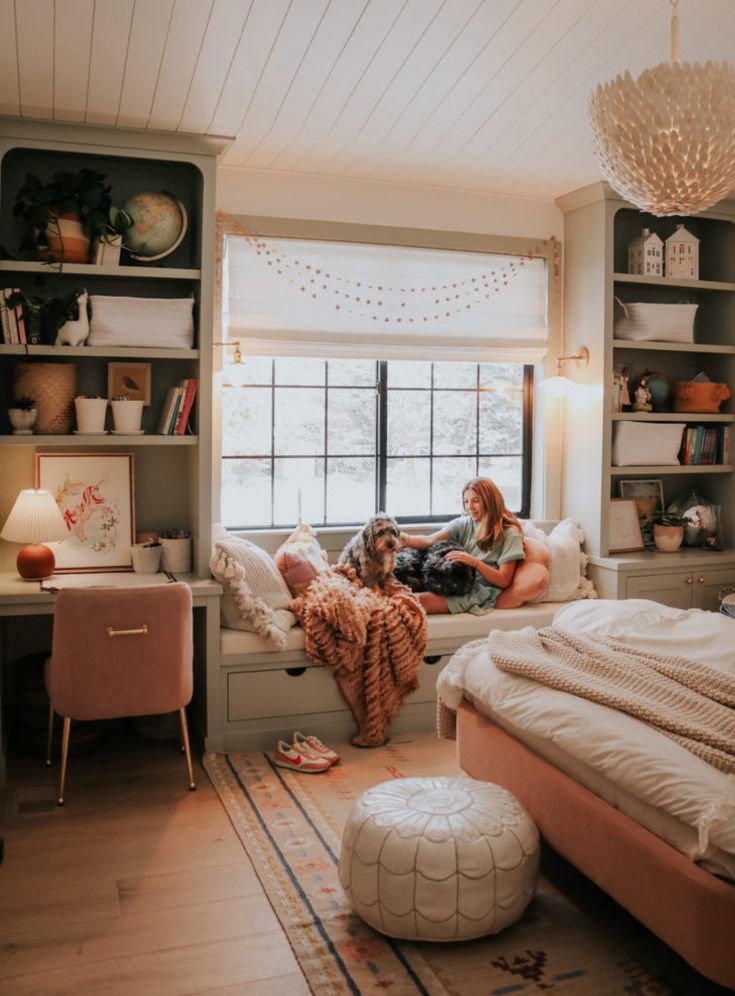 a woman sitting on a bed with her dog in front of the window and bookshelves