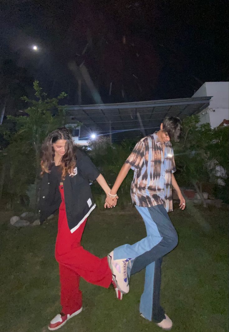 two people holding hands and dancing in the grass at night with lights on behind them