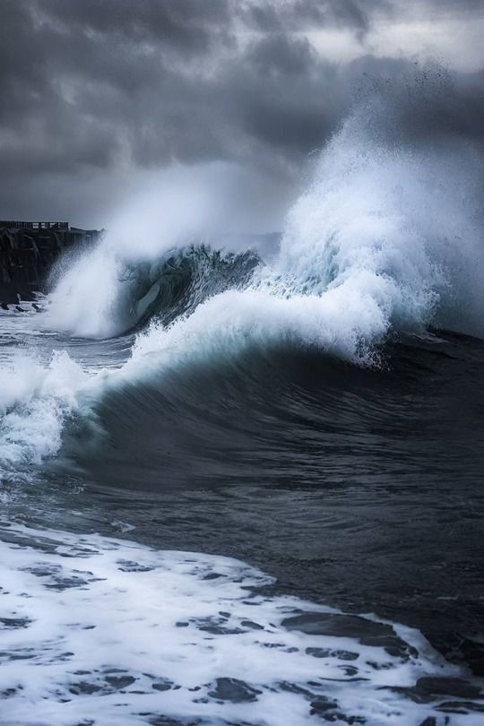 a large wave crashing into the ocean on a cloudy day with dark clouds above it