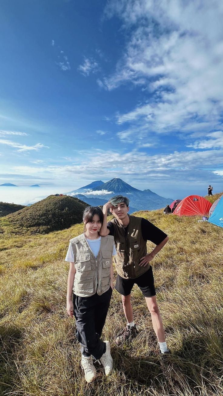 two people standing next to each other on top of a grass covered hill with tents in the background