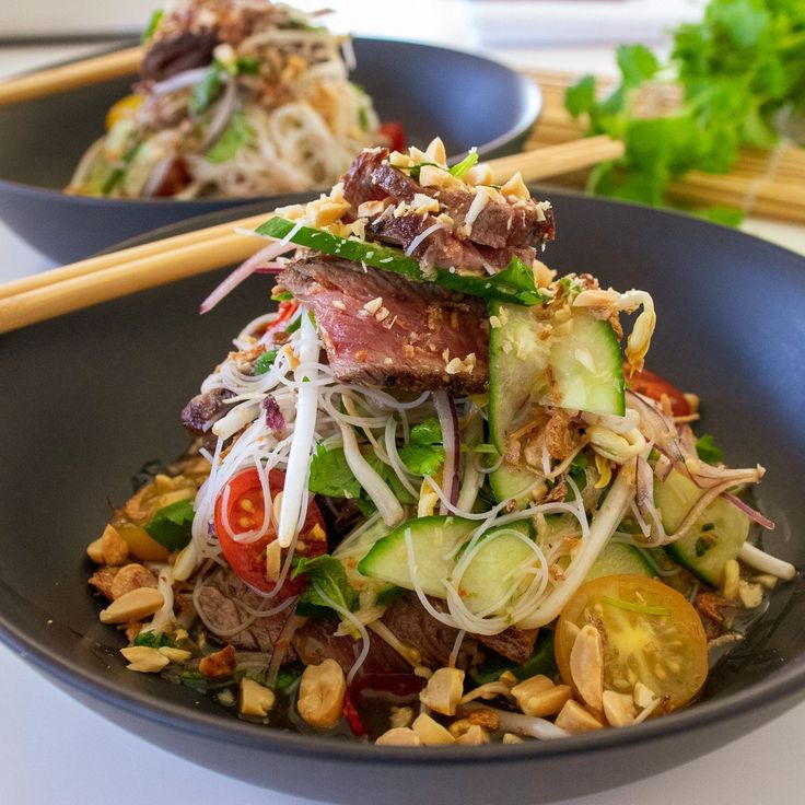 two black bowls filled with food and chopsticks on top of a white table