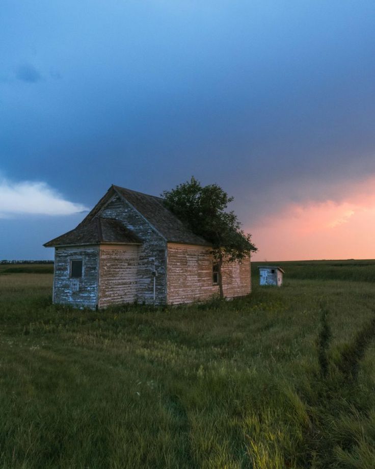 an old run down house in the middle of a grassy field at sunset with storm clouds overhead