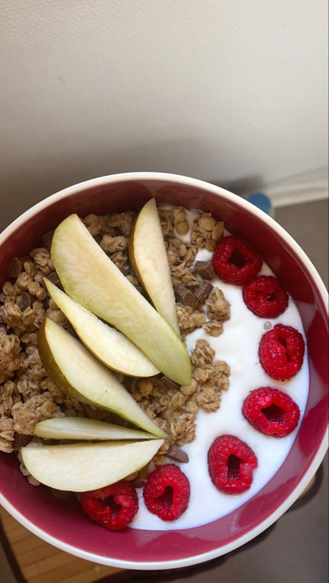 a bowl filled with granola, apples and raspberries on top of a table