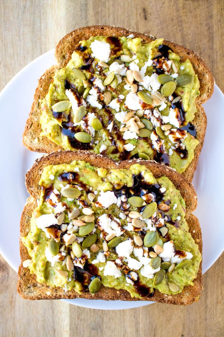 two pieces of bread with avocado and nuts on them sitting on a white plate