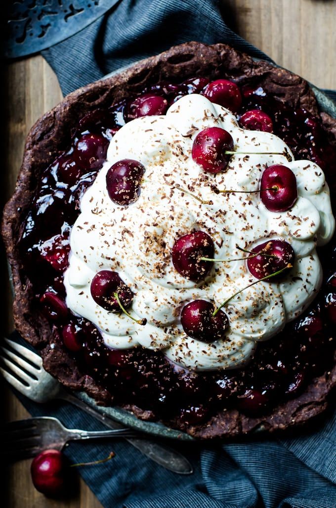 a pie topped with whipped cream and cherries on top of a wooden table next to two forks